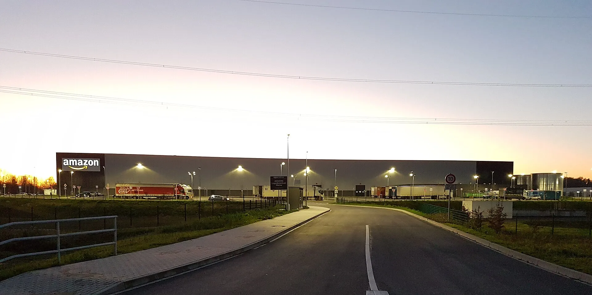 Un edifici de magatzem molt llarg i baix, gris, amb el rètol «amazon» il·luminat a l'esquerra, camions aparcats al davant i fanals encesos, al capvespre, vist des d'una carretera d'accés buida amb tanques a banda i banda.