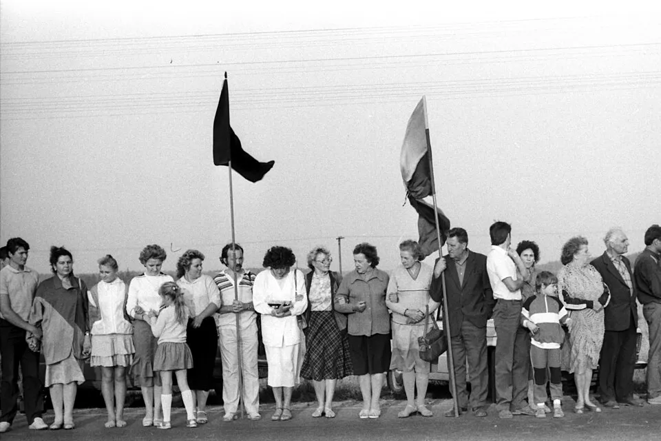 Fotografia en blanc i negre d'una filera de persones agafades de les mans a la vora d'una carretera, homes, dones i criatures amb roba d'estiu, dues banderes en pals, la primera negra i la segona de tres franges; línies elèctriques al fons i un cel gris.