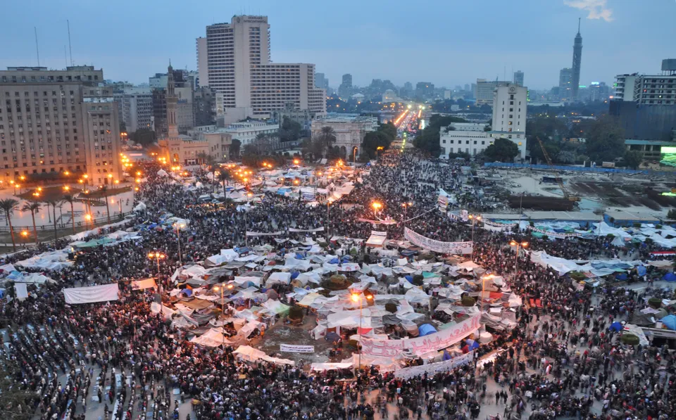 Vista des de dalt de la plaça Tahrir del Caire al capvespre, plena de gent fins als carrers laterals: al mig, una rotonda coberta de tendes i lones blanques i blaves; pancartes blanques amb lletres en àrab; a l'esquerra un gran edifici administratiu, al fons gratacels amb els llums encesos i a la dreta la torre del Caire.