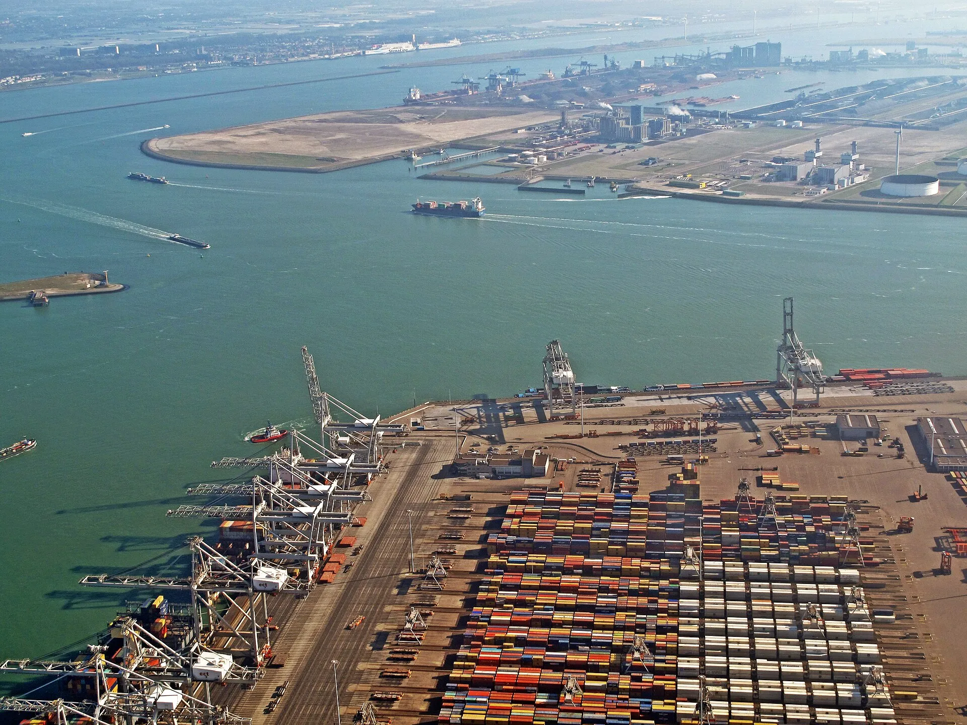 Vista des de dalt d'una esplanada immensa de contenidors de colors apilats en fileres, grues blaves al fons i, més enllà, el mar i un cel gris; cap persona a la vista.