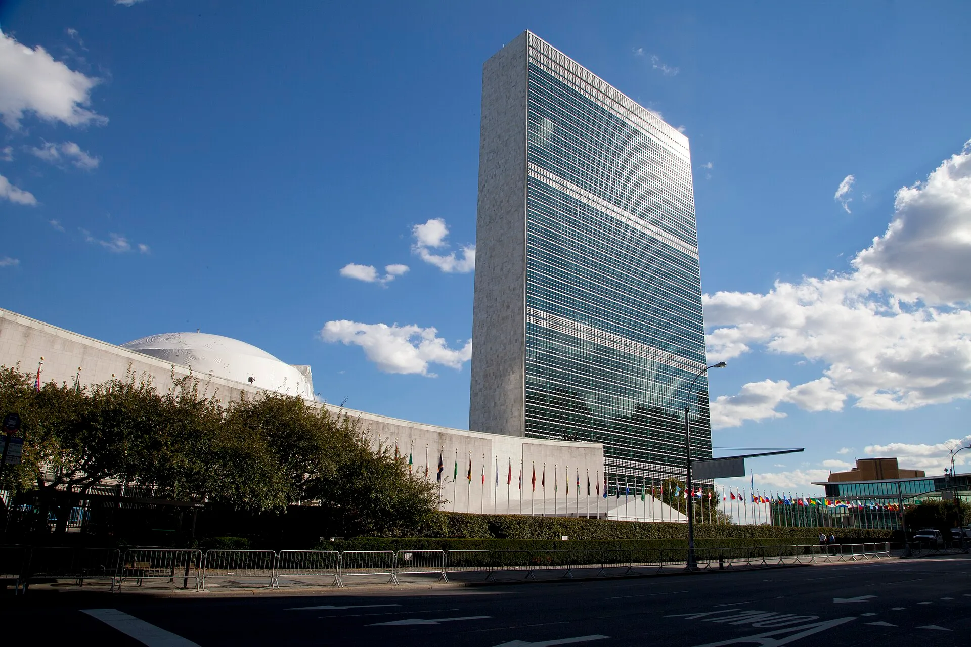La seu de les Nacions Unides a Nova York sota un cel blau amb núvols: la torre de vidre de la Secretaria a la dreta, la cúpula blanca de l'Assemblea General a l'esquerra darrere dels arbres, i una filera de banderes de tots els països davant de l'edifici baix, amb tanques al carrer.