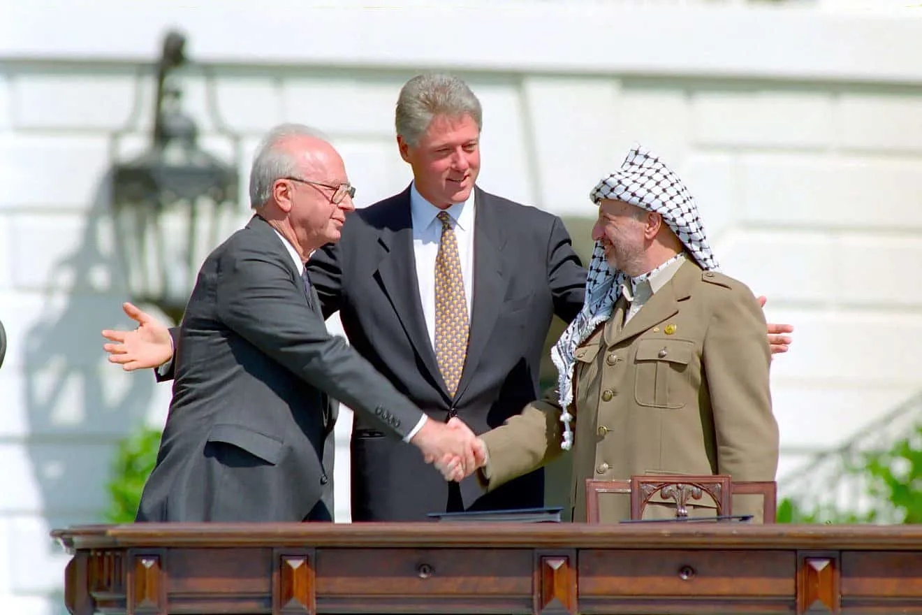 Tres homes als jardins de la Casa Blanca: al mig, Bill Clinton amb els braços oberts; a l'esquerra, Yitzhak Rabin amb vestit fosc; a la dreta, Yasser Arafat amb la kefia i uniforme; Rabin i Arafat es donen la mà davant d'un faristol.