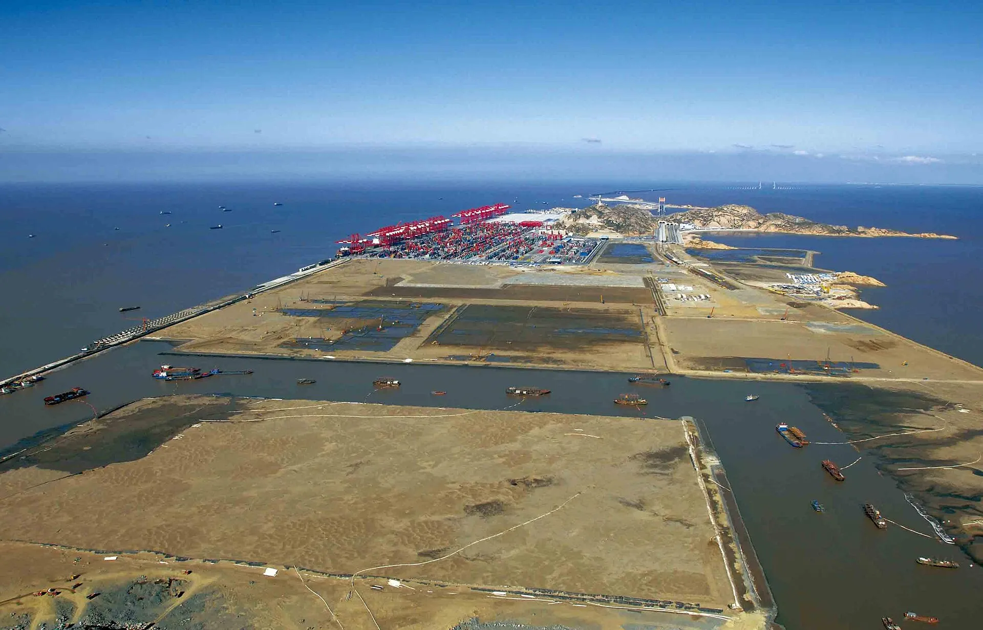Vista des de dalt d'un moll enorme amb fileres de grues vermelles i contenidors de colors apilats, un vaixell portacontenidors atracat a l'esquerra i el mar gris al fons.