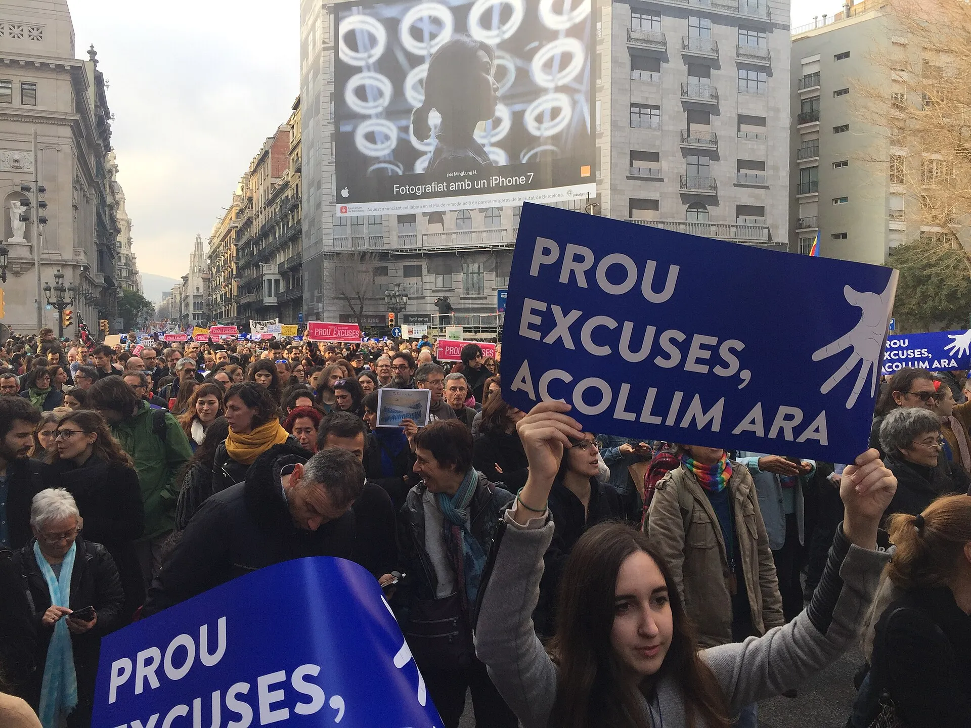 Una manifestació omple un carrer ample de Barcelona a la tarda; en primer pla, una noia aixeca una pancarta blava amb lletres blanques que diu «Prou excuses, acollim ara» i el dibuix d'una mà; al fons, una façana amb un cartell publicitari enorme i milers de persones fins on arriba la vista.