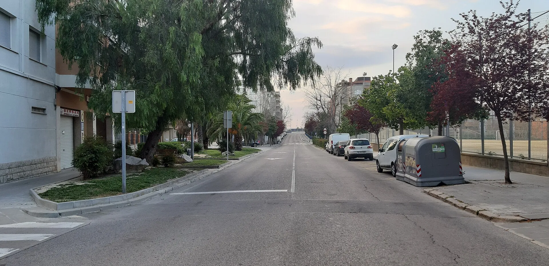 Un carrer ample i buit d'una ciutat mitjana a primera hora del matí: cap cotxe en marxa, alguns d'aparcats a la dreta, contenidors grisos, arbres verds i vermells a les voreres, un semàfor i una persiana de botiga abaixada a l'esquerra, i al fons la carretera que puja cap a uns blocs de pisos sota un cel clar.