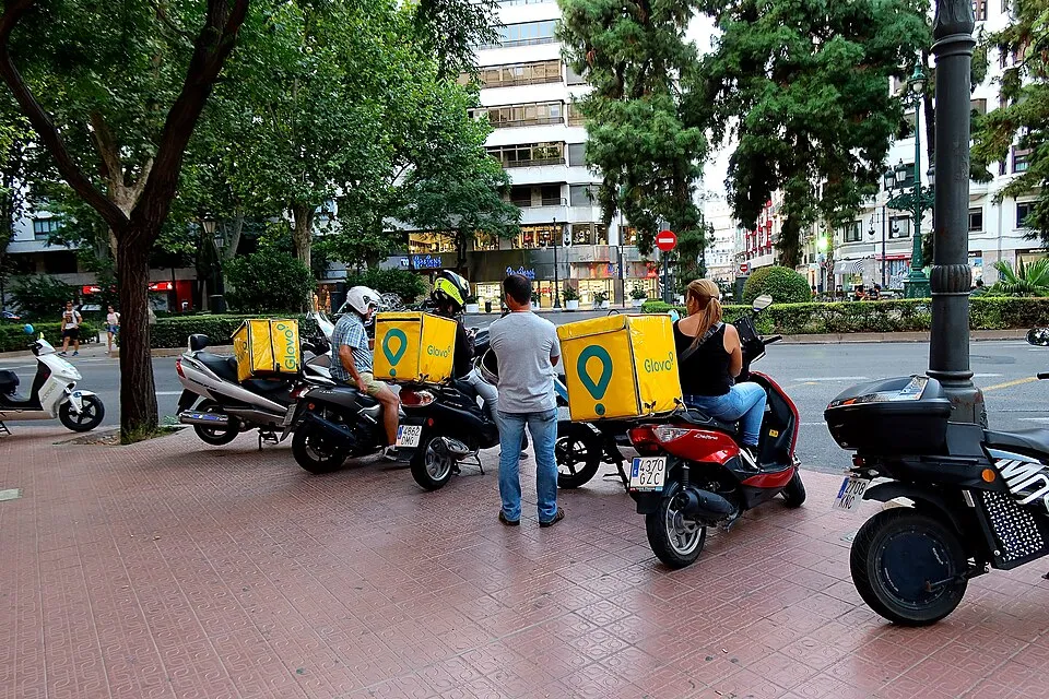 Una vorera de València amb quatre motos aparcades, tres amb caixes grogues de Glovo al darrere; dos repartidors amb casc asseguts a les motos, un home dret de cara a ells i una dona amb caixa groga a la moto de la dreta, davant d'una avinguda amb arbres i edificis.