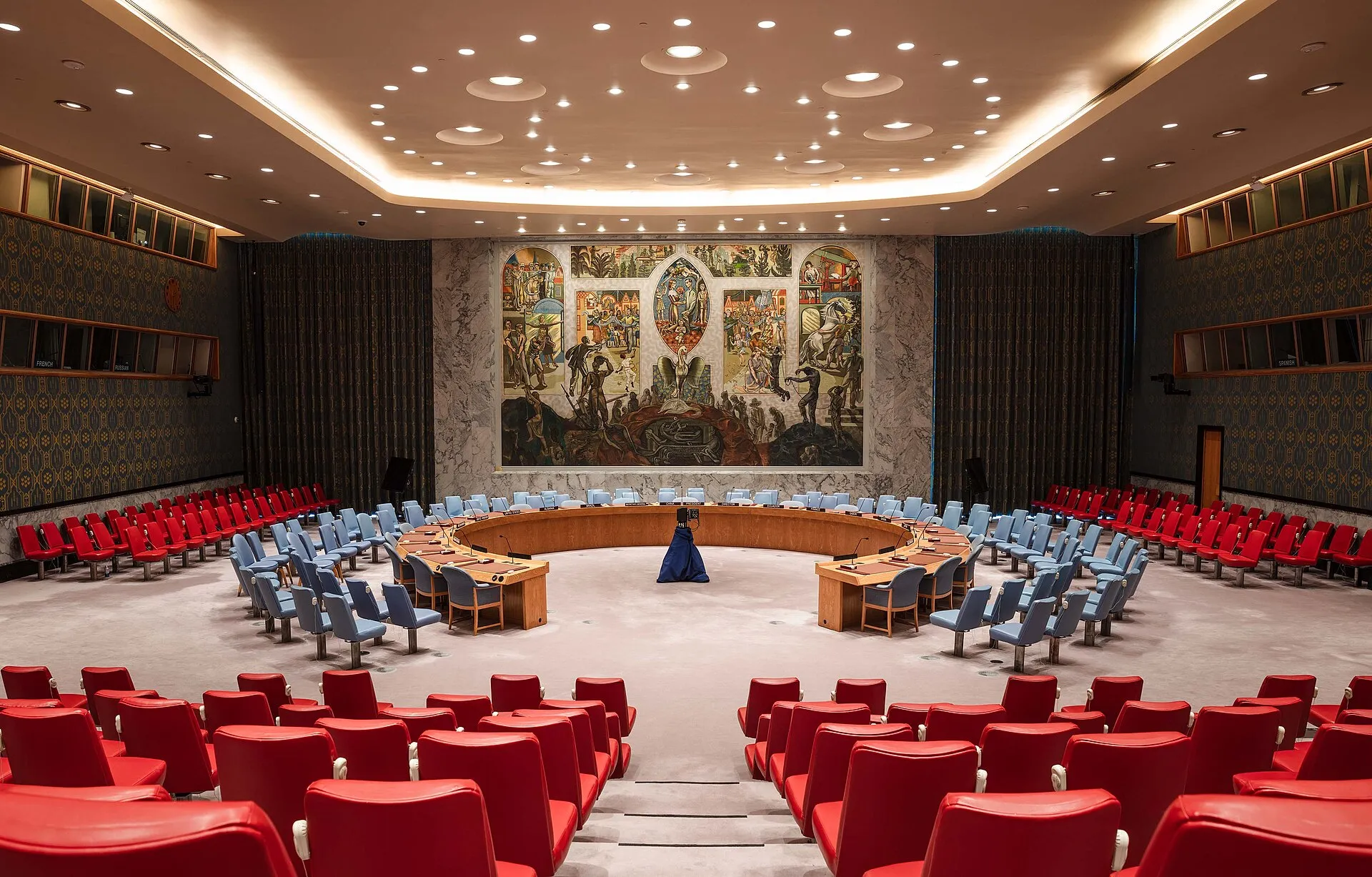 La sala del Consell de Seguretat de l'ONU buida: la taula en ferradura amb cadires blaves al voltant, fileres de butaques vermelles a primer pla i als costats, i al fons un gran mural amb figures que surten de la foscor cap a la llum, sota un sostre amb llums rodones.