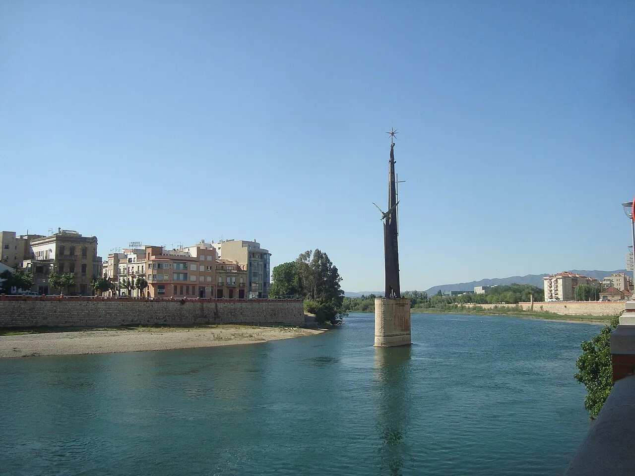 Un monument de ferro alt i estret que s'aixeca sobre un pilar al mig del riu Ebre, a Tortosa, amb la ciutat a la riba.