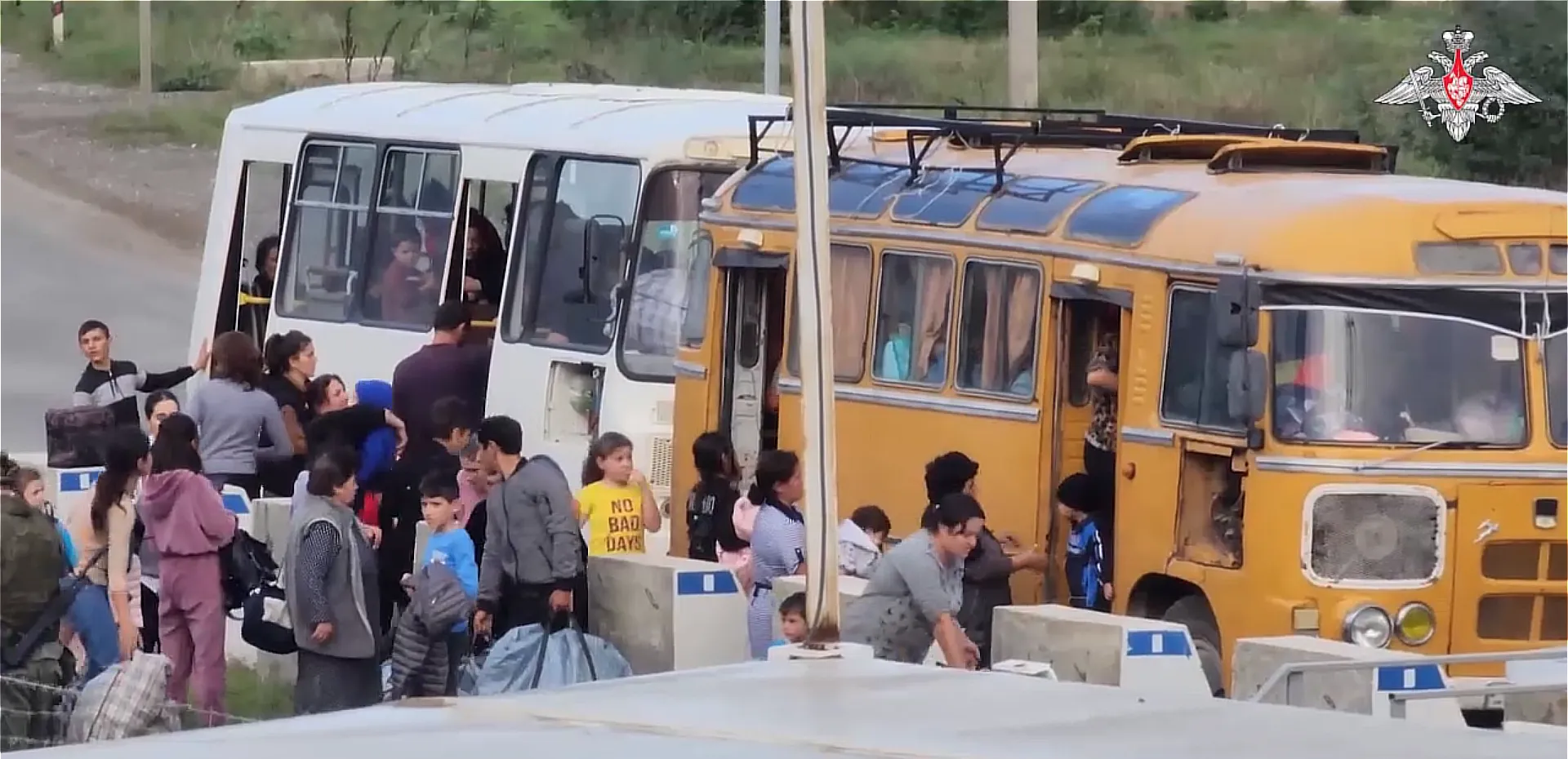 Fotograma d'un vídeo: dos autobusos vells, un de blanc i un de groc, aturats en una carretera, amb desenes de persones al voltant, dones, homes i criatures amb bosses i farcells, algunes pujant per la porta, i blocs de formigó al primer pla.
