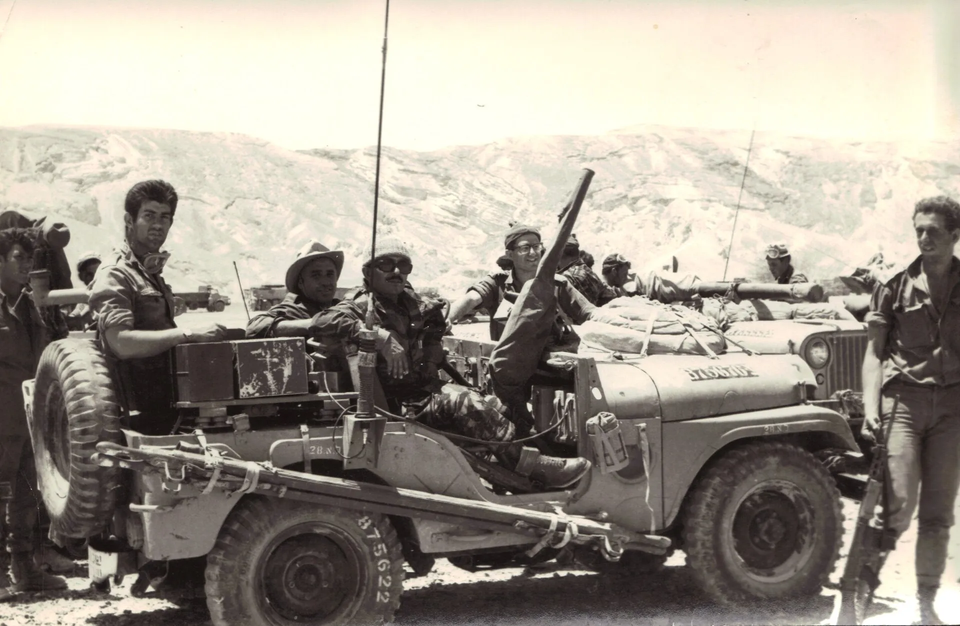 Fotografia en blanc i negre, de to sèpia, d'un jeep militar aturat en un paisatge de muntanyes àrides: cinc soldats amb uniforme i gorres, un amb ulleres de sol i bigoti al volant, un altre dret al costat amb un fusell, i una antena llarga que puja del vehicle.
