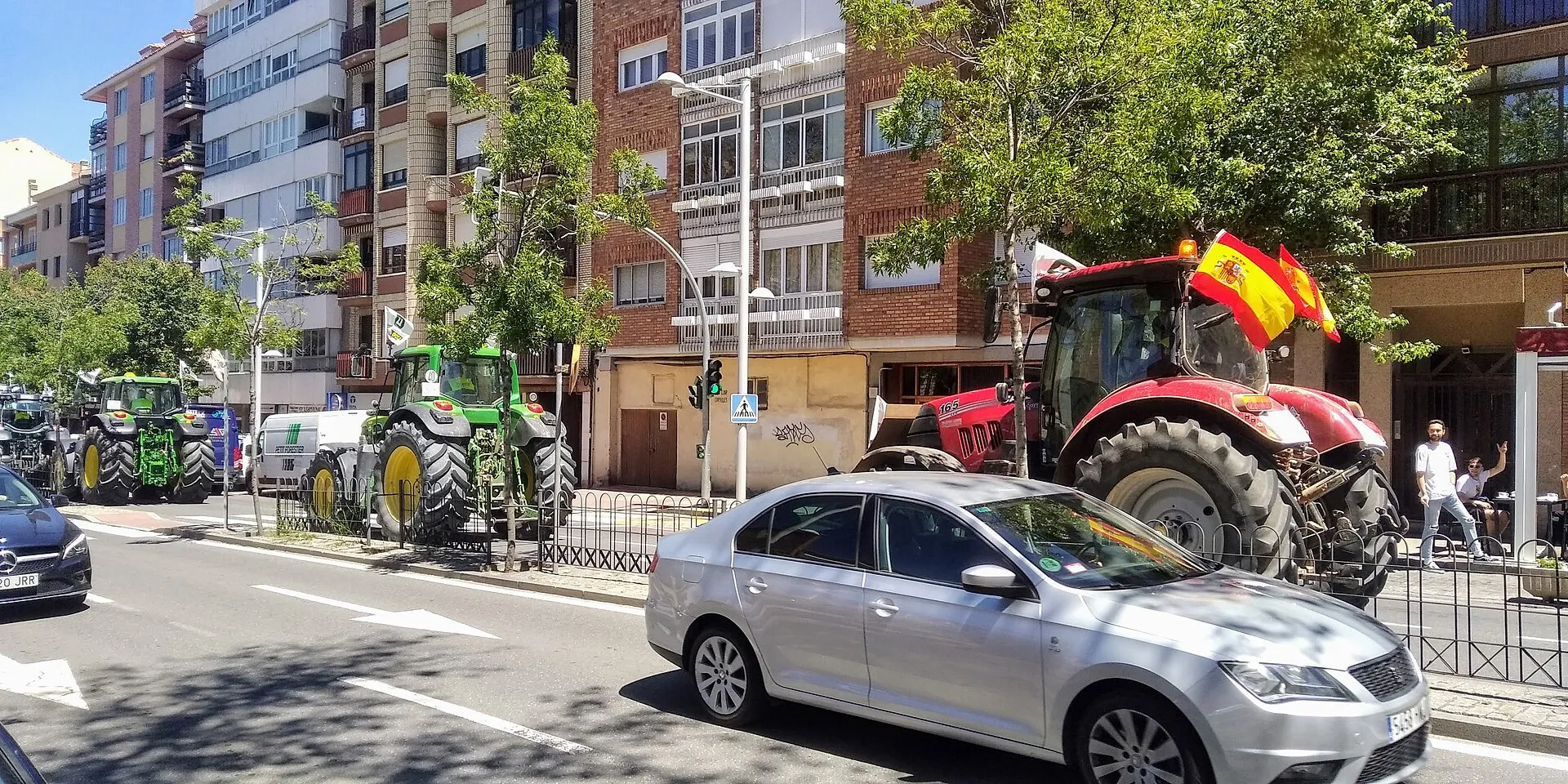 Una filera de tractors verds i vermells circula per un carrer de ciutat entre edificis de maons i arbres, un d'ells amb una bandera espanyola, mentre un cotxe gris passa per davant i dues persones miren des de la vorera.