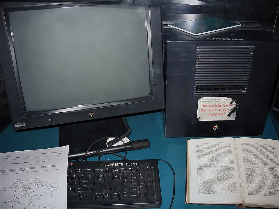 Un ordinador NeXT negre en forma de cub sobre una taula d'exposició, amb un monitor negre a l'esquerra, un teclat amb l'etiqueta «Propriété CERN», un adhesiu vermell al cub que diu «This machine is a server. DO NOT POWER IT DOWN!!», un llibre obert a la dreta i, a l'esquerra, un full imprès amb el títol «Information Management: A Proposal» i un diagrama de cercles i fletxes.