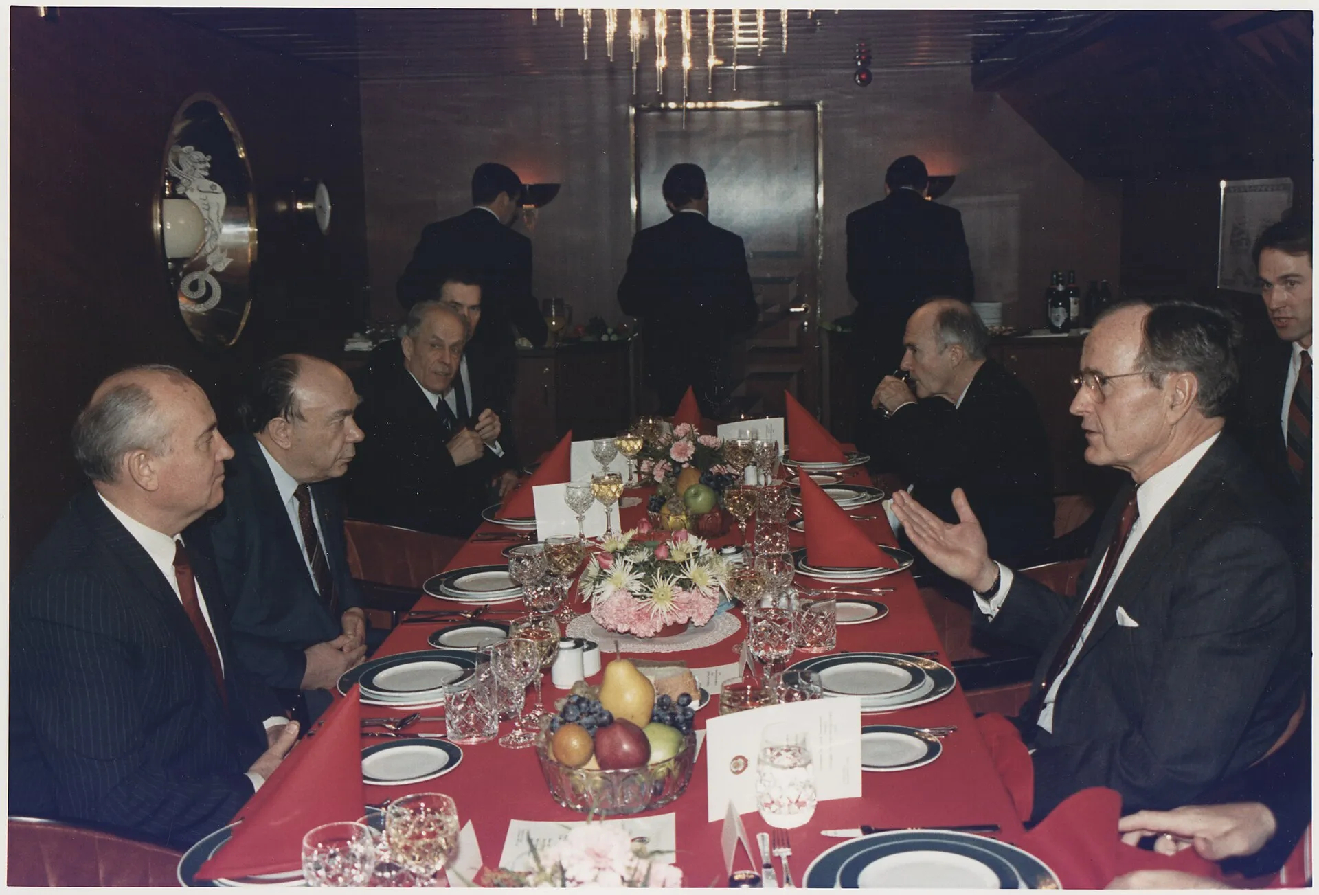 Una taula llarga en un menjador de vaixell, amb homes de vestit fosc asseguts a banda i banda; al centre, George Bush i Mikhaïl Gorbatxov, un davant de l'altre, parlen mentre els altres escolten; copes i plats a la taula i finestres rodones al fons.