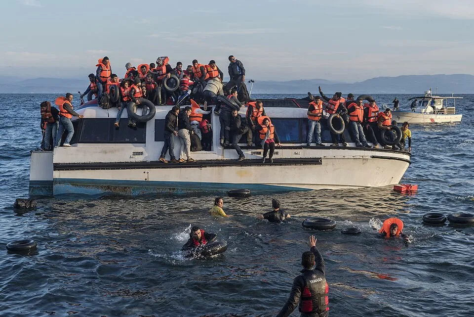 Una embarcació blanca petita atapeïda de persones amb armilles salvavides taronges, algunes de dretes al sostre, arriba a la costa; a l'aigua, mitja dotzena de persones neden o s'aguanten a flotadors negres, i una d'esquena amb vestit de neoprè aixeca la mà cap a la barca; al fons, una llanxa de vigilància i la costa de Turquia.