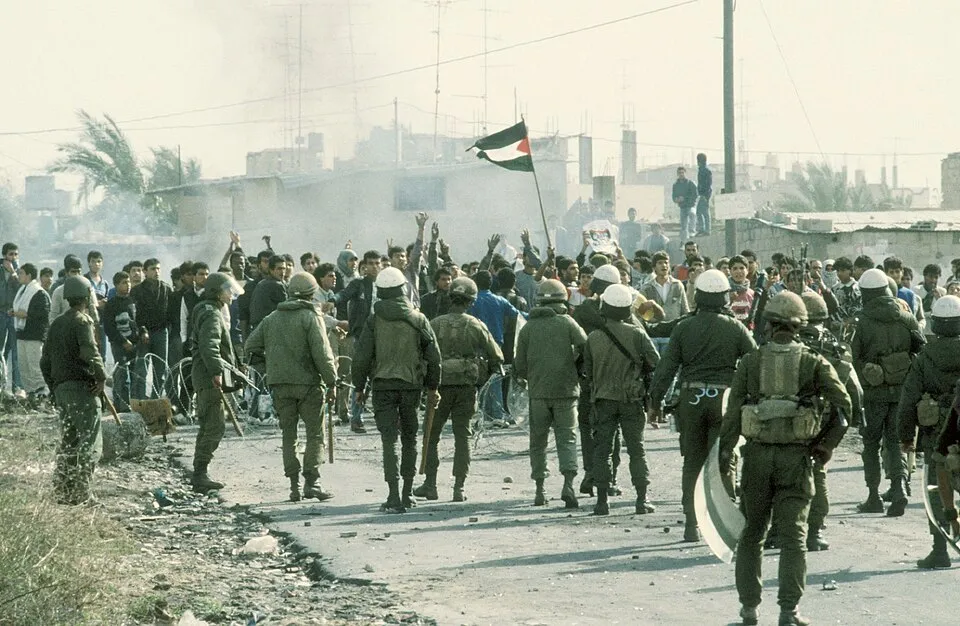 Fotografia en color d'un carrer de terra a Gaza: una filera de soldats israelians amb cascs blancs i uniforme verd, d'esquena, amb escuts i porres, davant d'una multitud de joves palestins que aixequen els braços, una bandera palestina al mig, fum al fons i cases baixes amb antenes.