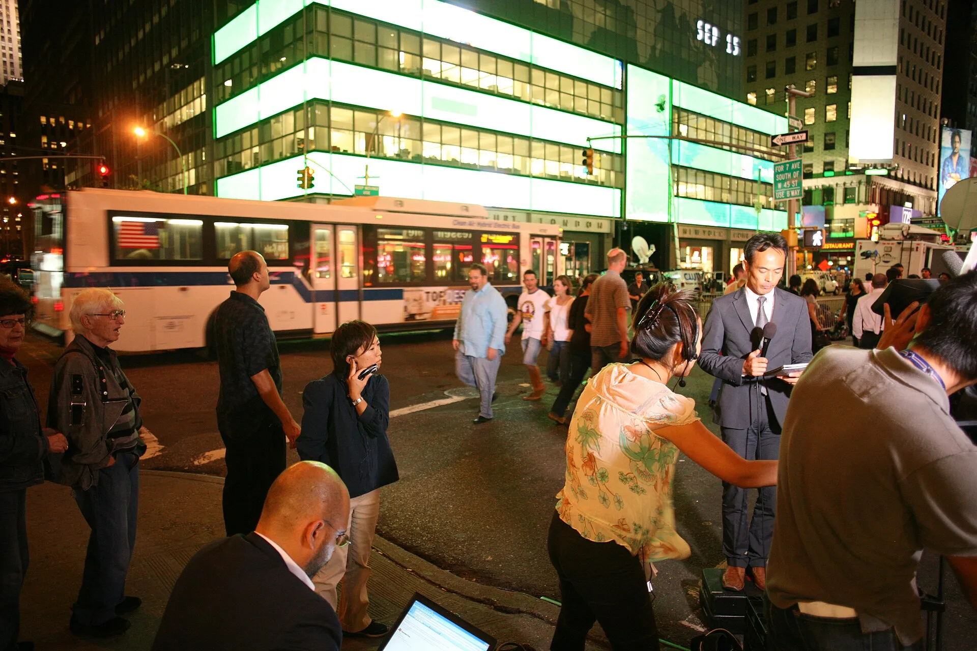 Un carrer de Nova York de nit davant d'un edifici de vidre amb franges il·luminades de verd; un autobús blanc passa desenfocat, gent parada a la vorera mirant, i a primer pla un periodista amb micròfon, una càmera i un home amb un portàtil.
