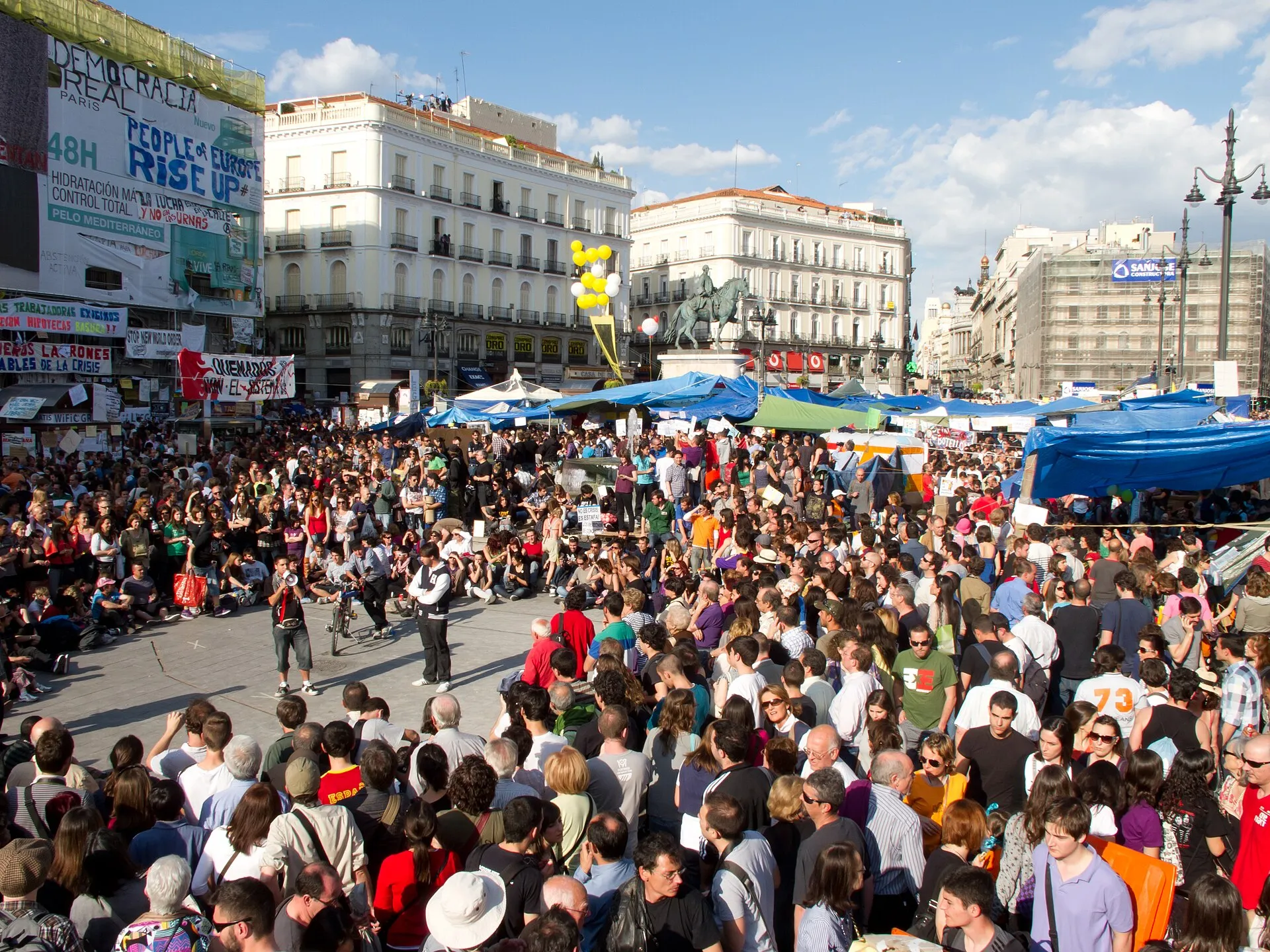 La Puerta del Sol de Madrid plena de gent a la tarda, amb lones blaves de l'acampada al mig, globus grocs, pancartes penjades de les façanes (una diu «Democracia real ya» i una altra «People of Europe rise up») i l'estàtua eqüestre al fons entre els edificis blancs.