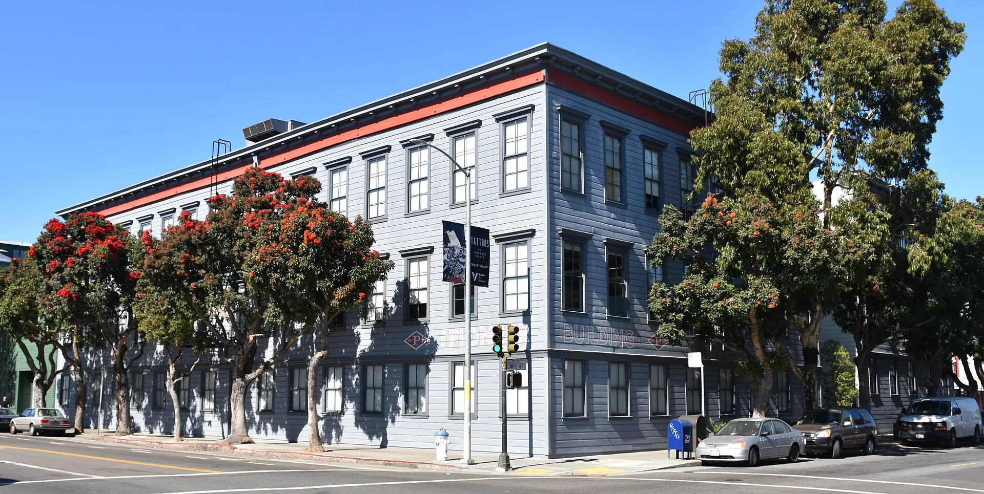 Un edifici de fusta gris de tres plantes amb cornisa vermella en una cantonada de San Francisco, amb finestres de guillotina, arbres de flor vermella a les dues voreres, un semàfor, cotxes aparcats i un cel blau sense núvols.